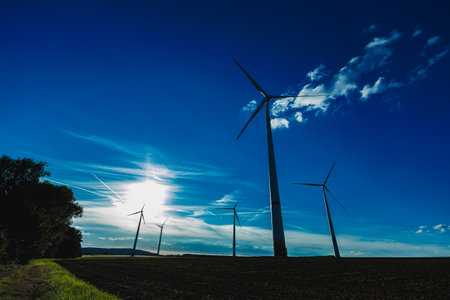 Wind turbines rise above a green landscape under a clear blue sky, capturing the sun's rays as they turn slowly in the wind during a bright day in the countryside.の写真素材