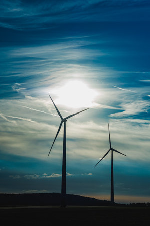 Two wind turbines tower in the foreground while the sun illuminates the clouds in a vibrant sky. This serene scene highlights the importance of renewable energy in nature.の写真素材
