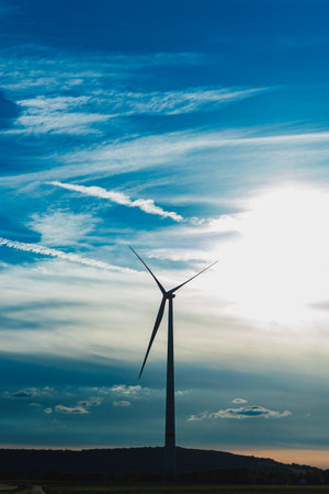 A wind turbine is silhouetted against a bright sun and colorful clouds. The scene captures the beauty of renewable energy in nature during late afternoon.の写真素材