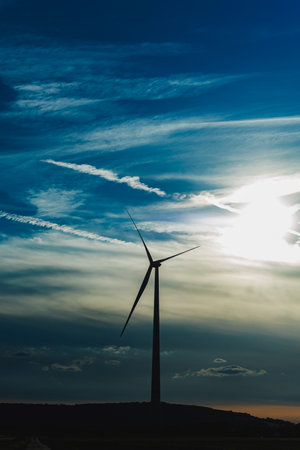 A wind turbine reaches high into the sky as the sun sets, casting a warm glow against a backdrop of swirling clouds and crisscrossing airplane trails. The scene highlights renewable energy.の写真素材