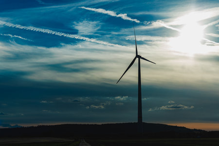 A tall wind turbine stands proudly on a hill, its silhouette stark against the colorful sunset sky filled with soft, wispy clouds. The scene captures the beauty of renewable energy and nature.の写真素材