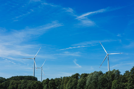 Tall wind turbines turn slowly against a bright blue sky, surrounded by lush green trees, showcasing...