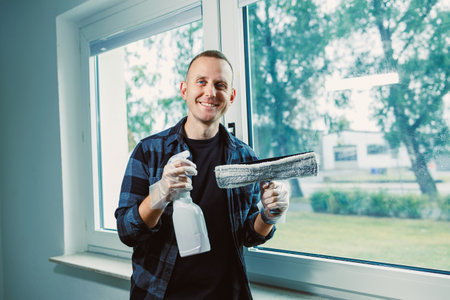 A cheerful man stands by a large window, holding a spray bottle and a window cleaning tool, ready to...