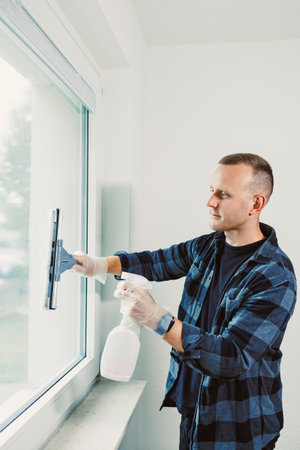 A man dressed in a plaid shirt and gloves meticulously cleans a large window, using a spray bottle...