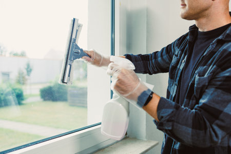 A person is focused on cleaning a window using a squeegee and spray cleaner, ensuring sunlight streams into the room while revealing a green outdoor landscape.の写真素材