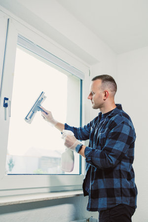 A man wearing gloves diligently cleans a window in a contemporary room filled with natural light,...