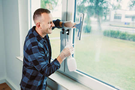 A cheerful man in a blue checkered shirt cleans windows with a squeegee and spray bottle, enjoying the sunny afternoon in a bright, modern home. Freshness fills the air as he smiles.の写真素材