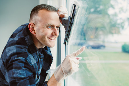 A cheerful man wearing gloves and a checkered shirt cleans a window, intent on making it sparkle. Sunlight floods the room, enhancing the positive vibe of the moment.の写真素材