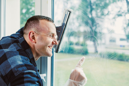 A person smiles while cleaning a window, enjoying the fresh view outside. Sunlight illuminates the scene, creating a cheerful and productive atmosphere.の写真素材