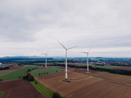 Three wind turbines rise above green fields and brown patches, set against a moody sky. The scene highlights clean energy's role in peaceful rural life. Nature and technology unite beautifully.の写真素材