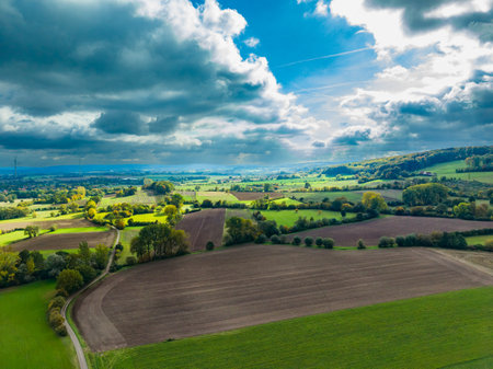 Vibrant green fields stretch across the landscape, framed by dark clouds and sunlight breaking through. A winding road leads through the serene countryside, showing nature's beauty.の写真素材