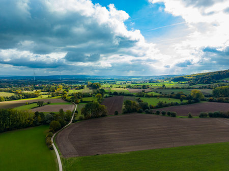 Vast green fields and patches of earthy soil stretch out beneath a dynamic sky filled with clouds. A winding road cuts through the landscape, showing rural tranquility during daytime.の写真素材