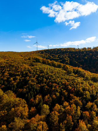 Colorful autumn foliage blankets rolling hills as tall wind turbines stand against the clear blue sky, creating a striking contrast with nature's artistry.の写真素材