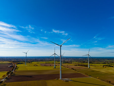 Wind turbines stand tall in a vast green field under a bright blue sky. The landscape stretches far,...