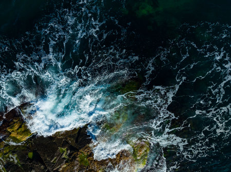 Bright sunlight reflects off the ocean as waves vigorously crash against the rocky coastline, creating foamy splashes and revealing the beauty of water meeting stone.の写真素材
