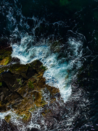 Waves surge powerfully over dark stones along a secluded shore, reflecting hues of orange and blue as the sun sets in the background, creating a serene and dynamic coastal scene.の写真素材