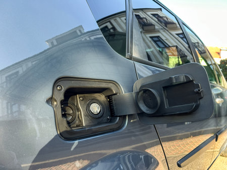 A person connects the charging cable to an electric vehicle in a bustling urban area. The sun casts...