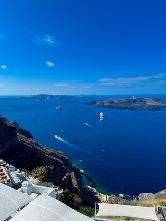 Glittering waters reflect the sun as boats navigate the serene sea around Santorini's volcanic islands. The sky is a vibrant blue without a cloud in sight.の写真素材