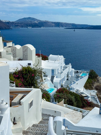 A picturesque scene unfolds with elegant white structures and turquoise pools lining the hillside, set against the deep blue sea and majestic mountains on a bright day.の写真素材