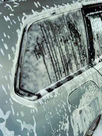 Soap suds cover a shiny silver car, creating a playful scene as water streams down its side, showcasing a lively afternoon of car cleaning in a sunny environment.の写真素材