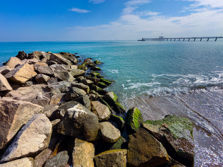 Under a clear blue sky, the shoreline features sturdy rocks washed by calm waves, revealing a peaceful seaside scene near a long fishing pier in the distance.の写真素材