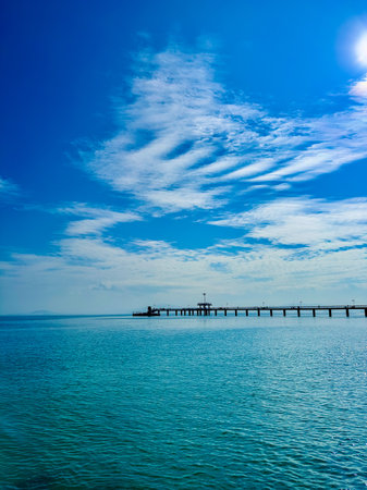 Bright sunlight reflects off calm waters near a peaceful pier, where a small boat moves gracefully. The sky is a stunning blue, dotted with wispy clouds.の写真素材