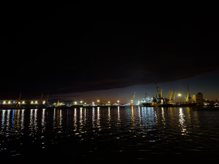 Harbor scene comes alive at night, featuring bright lights illuminating the waterfront and casting shimmering reflections on calm water, creating a serene urban atmosphere.の写真素材