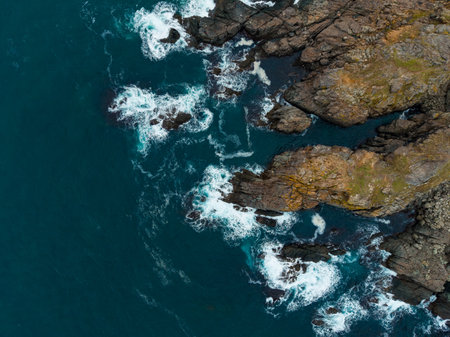 Rugged cliffs meet the ocean as waves crash against them. The water glimmers under a soft setting sun, creating a peaceful yet powerful scene in nature's embrace.の写真素材