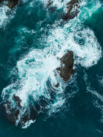 Waves surge and swirl around rugged rocks, creating a stunning display of white foam against deep...