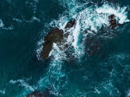 Dark rocks emerge from the vibrant blue sea as frothy waves crash around them, creating a serene and magnificent coastal scene under a clear sky.の写真素材
