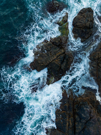 Powerful waves surge and crash against dark, jagged rocks at a beautiful coastal location during early morning. The tide reveals the mesmerizing movement of water.の写真素材