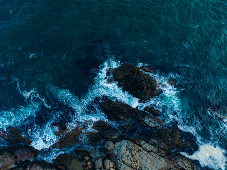 Bright blue waves crash onto dark, jagged rocks along the coast, foaming and splashing in the warm sunlight of early afternoon, showing nature's raw beauty and power.の写真素材