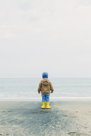 A young child stands at the edge of the beach, dressed in yellow boots and a cozy jacket. The cloudy sky contrasts with the tranquil blue sea, creating a serene atmosphere for exploration.の写真素材