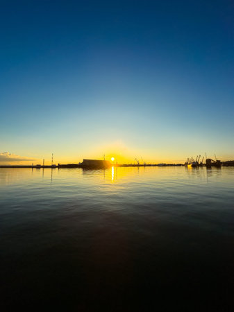 Sun rises above the water reflecting light as cranes stand in silhouette against a clear sky. The scene captures morning at a busy harbor with calm water.の写真素材