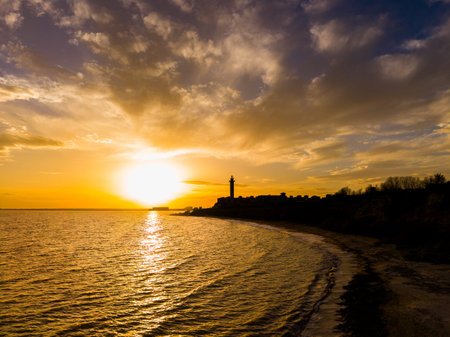 The sun sets over the water creating a bright glow in the sky while a lighthouse stands on the shore. Waves gently lap against the beach as clouds drift above.の写真素材