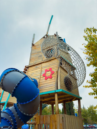 Children play at a playground featuring a large wooden structure with a slide and climbing areas. The sky is cloudy, and families enjoy the park around the playground.の写真素材