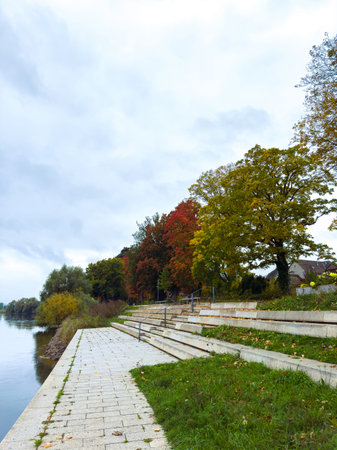 Stairs run along the river, with trees showing red and yellow leaves. The sky is cloudy, and the river reflects the surroundings. People can walk along the path.の写真素材