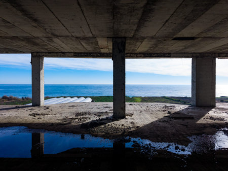 A person stands in an unfinished building looking out at the ocean. The scene shows the blue water, sky, and concrete pillars. The ground is wet, reflecting some light.の写真素材