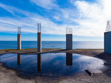 Concrete columns rise from a flat surface near the ocean. Water gathers in a pool, showing reflections of the sky. Waves roll silently in the background under a blue sky.の写真素材