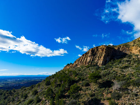 The scene shows a rocky hill surrounded by trees and shrubs. The sky is blue with some clouds. A valley stretches beneath the hill with fields and distant mountains.の写真素材