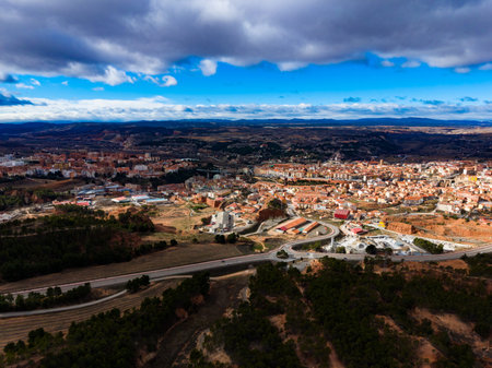 The scene captures Teruel city from above. The image shows a mix of buildings, roads, and natural terrain under a cloudy sky during daylight. The city is set against a backdrop of hills.の写真素材