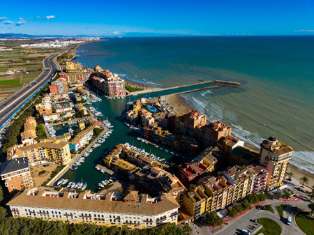 The scene shows a marina filled with boats surrounded by buildings. The coastline stretches into the distance under a bright sky and calm waters. Roads line the area, connecting to the town.の写真素材