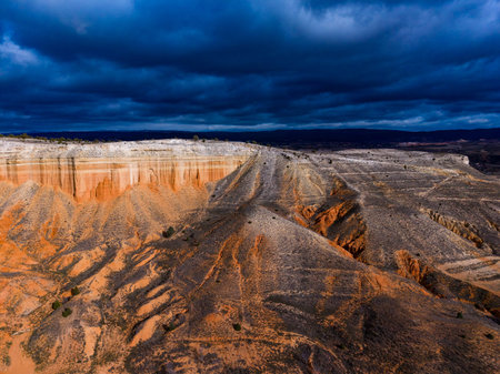 A canyon stretches across the land with steep walls. Dark clouds move overhead as shadows grow on the rocky ground. The scene shows signs of erosion and dry plants scattered around.の写真素材