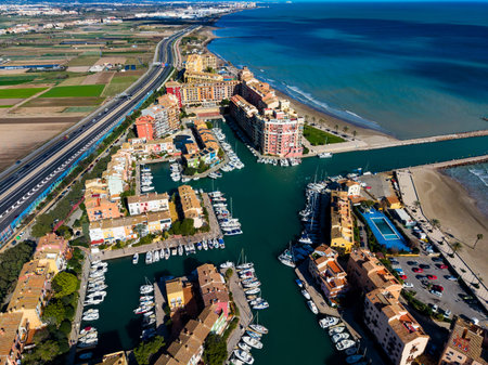 A coastal town features a marina filled with boats next to a highway and agricultural land. The scene shows buildings, water, and clear skies under bright sunlight.の写真素材