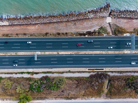 Cars travel on a highway beside a body of water with a rocky shore. Green plants line the edges of the road. The scene is bright and clear during the day.の写真素材
