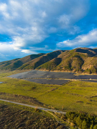 A view shows solar panels placed in a large area surrounded by grass and mountains. A road runs through the scene, with clouds in the sky above.の写真素材