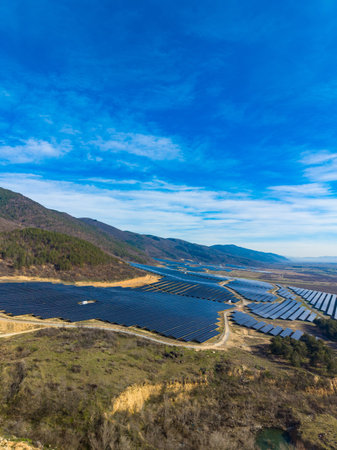 Solar panels stretch across the land near the mountains. The sky is clear with some clouds. The scene shows renewable energy development in a rural area in the afternoon.の写真素材