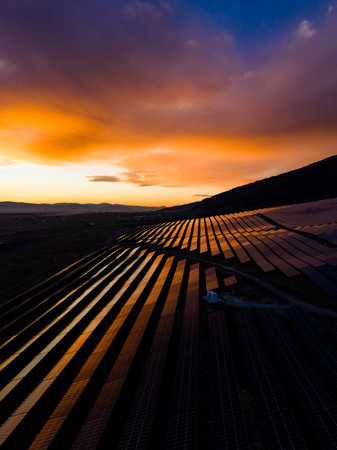 Sunlight hits the rows of solar panels at sunset. The panels reflect the colors of the sky. Hills are seen in the distance. The scene shows a clean energy setup in nature.の写真素材