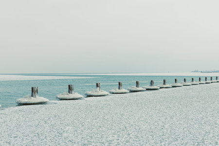 The scene shows a shoreline where water meets ice and snow. Wooden posts are visible along the water's edge. The sky is gray and overcast as winter continues.の写真素材