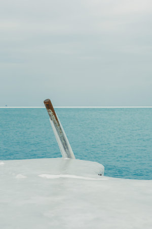 A boat's metal handle sticks out from a frozen surface while blue water stretches out in the background. Clouds cover the sky during this cold day at the lake.の写真素材